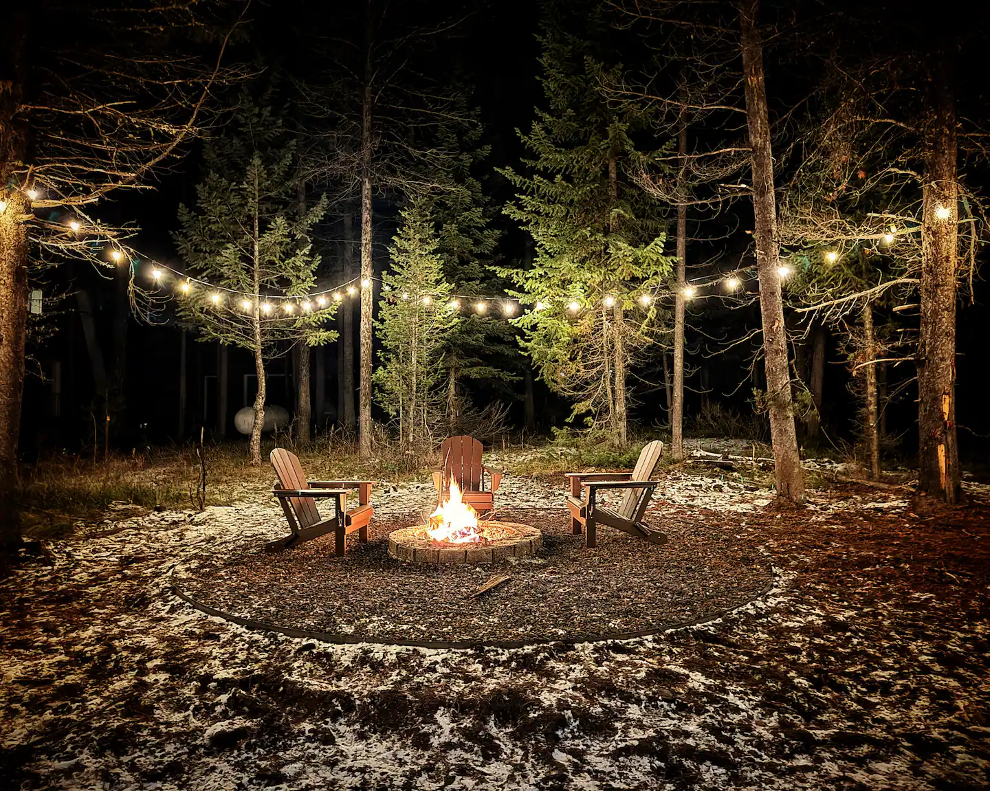 Firepit ringed by Adirondack chairs under string lights in the pines at night