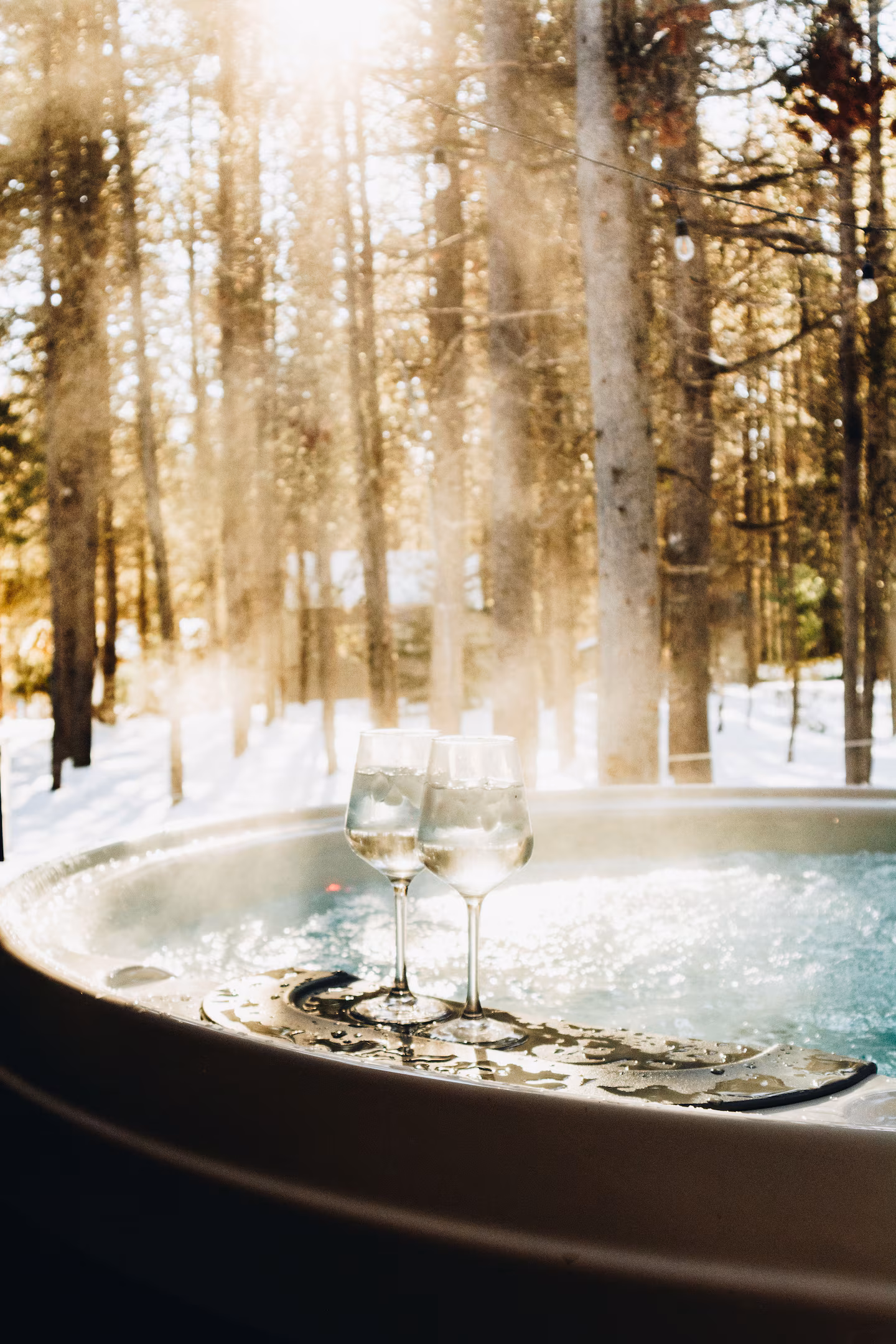 Steaming hot tub with two glasses on the rim, snow and sunlit pines beyond